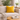 Mustard yellow Harris Tweed cushion on a beige sofa in a living room with a plant and coffee table in the background.