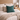 Peacock Harris Tweed cushion on a bed with pillows and a blanket, with a lamp and flowers on a nightstand.